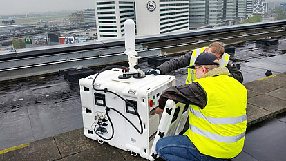Techniciens installant une unité de surveillance mobile Deux techniciens en gilets haute visibilité installent une unité de surveillance mobile sur un toit pour évaluer la qualité du signal et les performances du réseau.