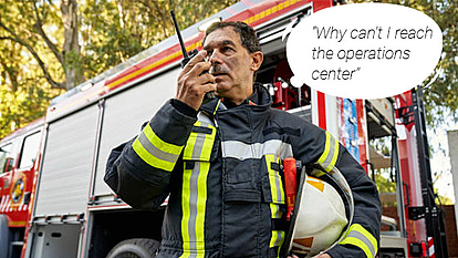 Firefighter using a radio in front of a fire truck, expressing difficulty in reaching the operations center. Represents the importance of reliable critical communication networks.
